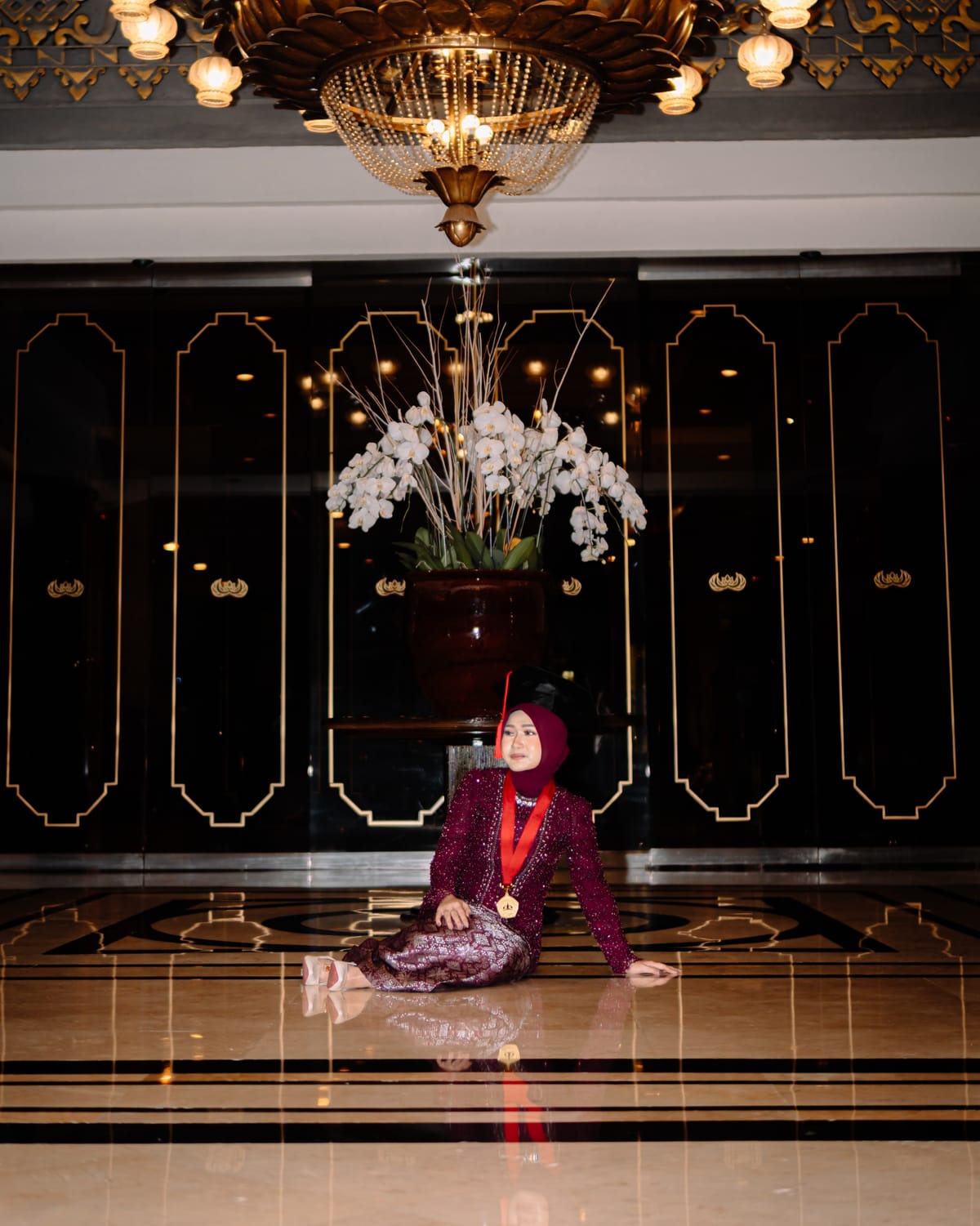 Wisudawati hukum berkebaya maroon, toga, dan medali alumni duduk di ballroom di bawah lampu gantung dengan anggrek putih