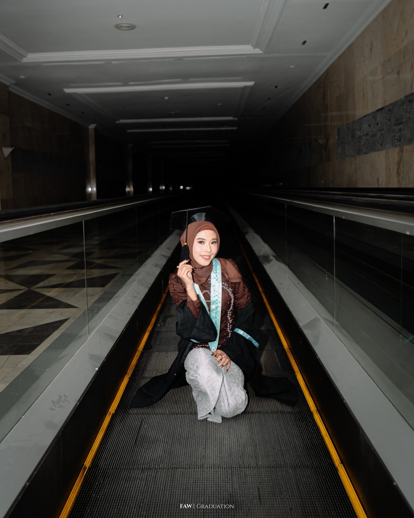 Wisudawati duduk di travelator dengan toganya saat sesi malam, garis kuning eskalator menyala