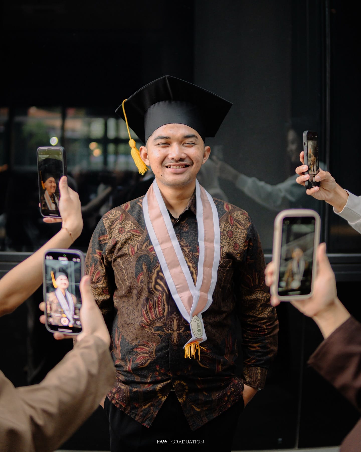 Wisudawan berkemeja batik dan toga dikelilingi tangan teman temannya yang memotret lewat ponsel