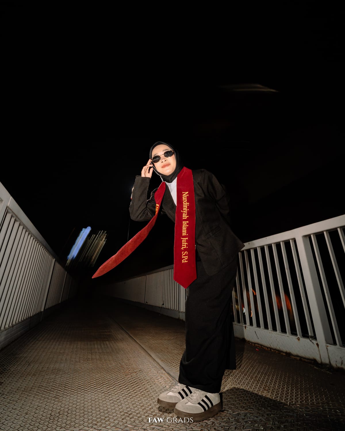 Wisuda malam di jembatan kota, wisudawati berhijab dengan toga hitam, selempang merah, kacamata hitam, dan sneakers, gaya urban