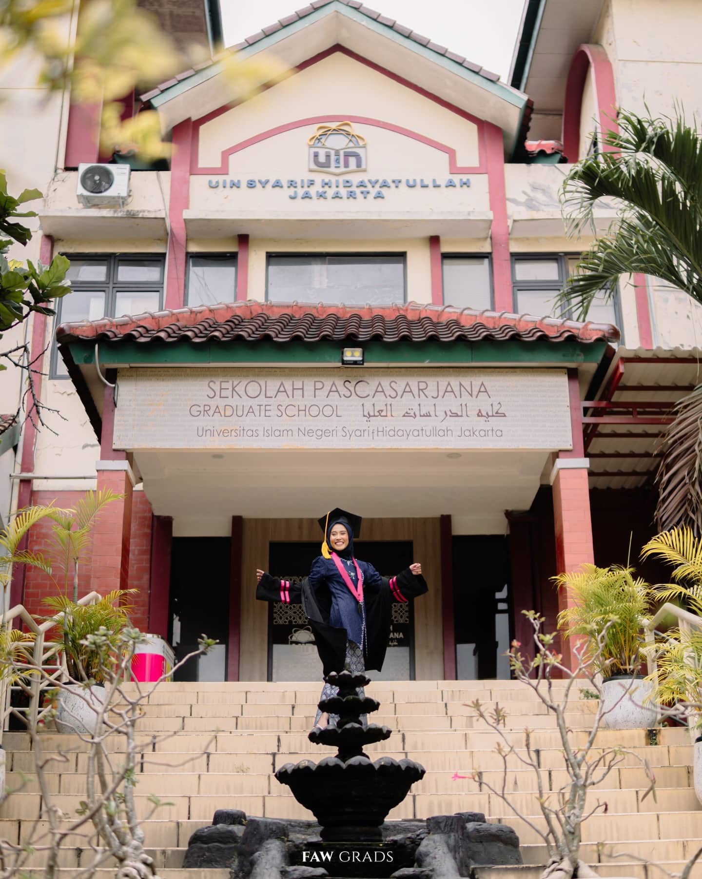 Wisudawati berhijab merentangkan tangan gembira di depan gedung Pascasarjana UIN Jakarta, toga hitam dengan hood magenta